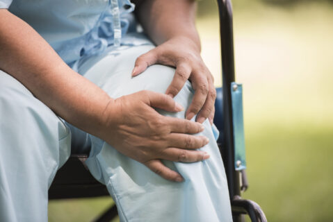 Elderly woman holding her painful knee while sitting in a wheelchair due to knee arthritis or chronic joint pain.
