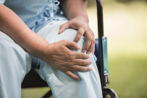 Elderly woman holding her painful knee while sitting in a wheelchair due to knee arthritis or chronic joint pain.